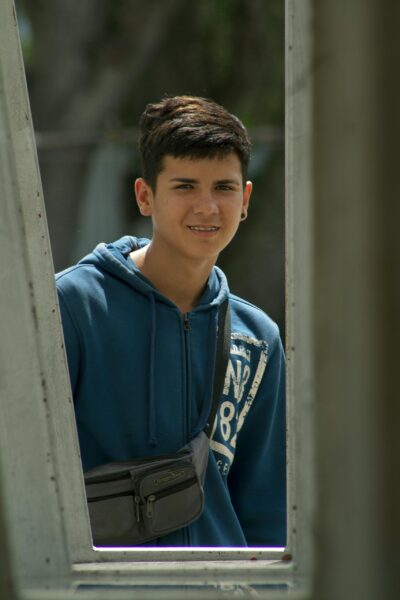 Hispanic teenage boy smiling in blue hoodie with sling bag outdoors. Bright day.